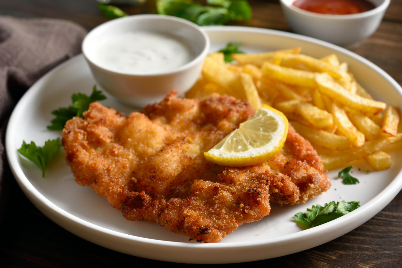 Close up view of breaded wiener schnitzel with potato fries and sauce on white plate over wooden table. Shallow depth of field.