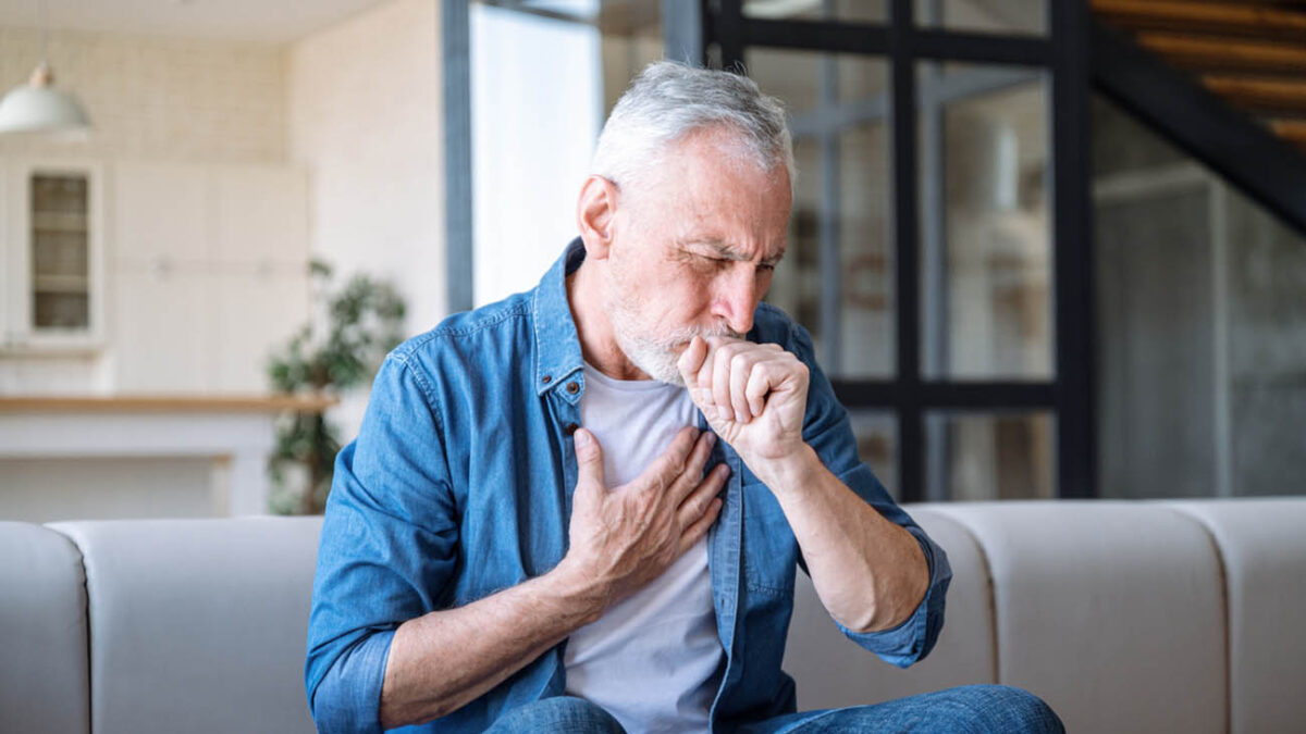 Senior man sitting on sofa at home and holding hand on chest. Male having asthma attack, difficulties with breathe, feeling severe pain or dyspnea