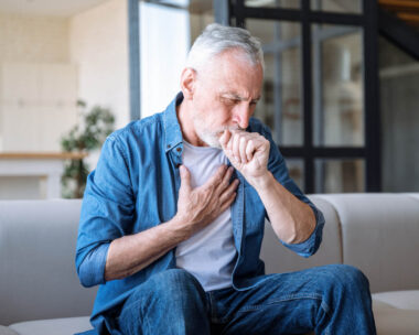 Senior man sitting on sofa at home and holding hand on chest. Male having asthma attack, difficulties with breathe, feeling severe pain or dyspnea