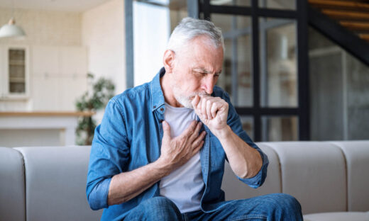 Senior man sitting on sofa at home and holding hand on chest. Male having asthma attack, difficulties with breathe, feeling severe pain or dyspnea