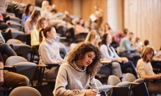 Female college student writing an exam during a class at lecture hall. Her classmate are in the background.