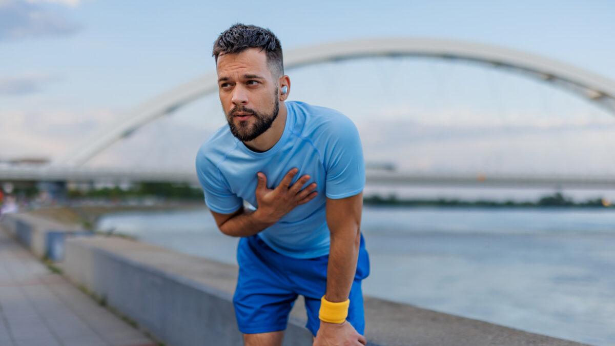 A man in a light blue shirt and blue shorts is exercising outdoors by the river, holding his chest and bending over, with a bridge in the background, conveying fatigue.