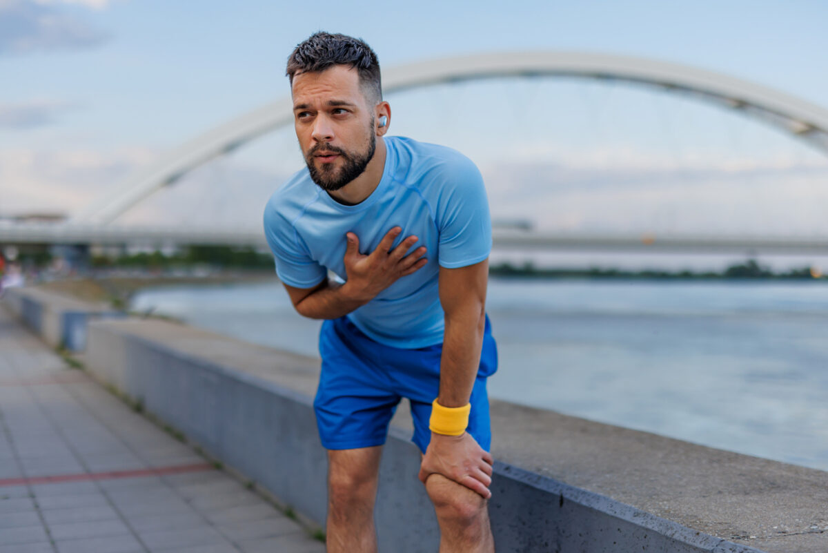 A man in a light blue shirt and blue shorts is exercising outdoors by the river, holding his chest and bending over, with a bridge in the background, conveying fatigue.