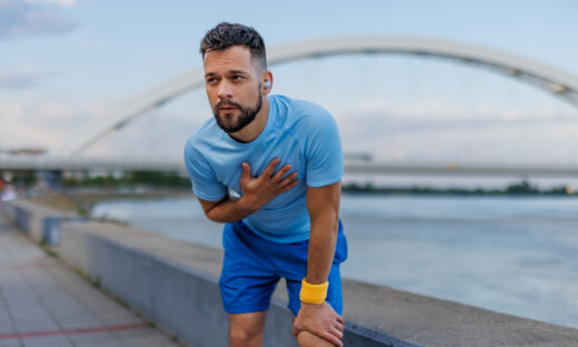 A man in a light blue shirt and blue shorts is exercising outdoors by the river, holding his chest and bending over, with a bridge in the background, conveying fatigue.