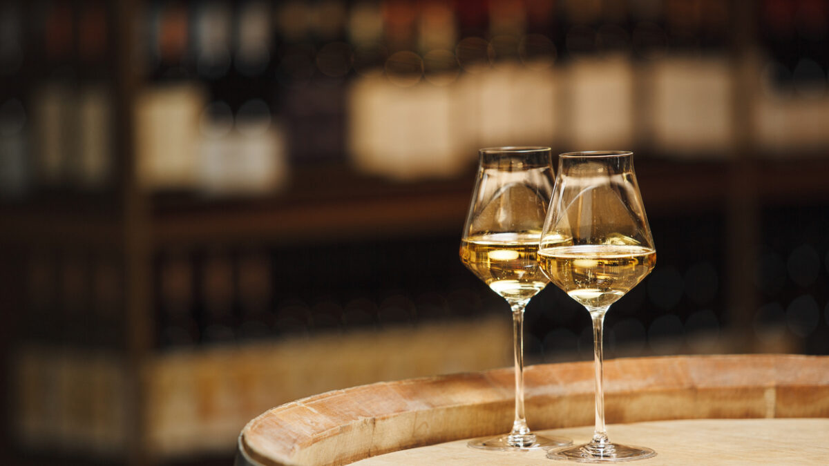 Two glasses of white wine sit on a wooden barrel in winery setting. The glasses are filled with clear, golden liquid and are partially illuminated by the ambient light against background is out of focus, showing the shelves of wine cellar.