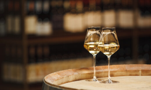 Two glasses of white wine sit on a wooden barrel in winery setting. The glasses are filled with clear, golden liquid and are partially illuminated by the ambient light against background is out of focus, showing the shelves of wine cellar.