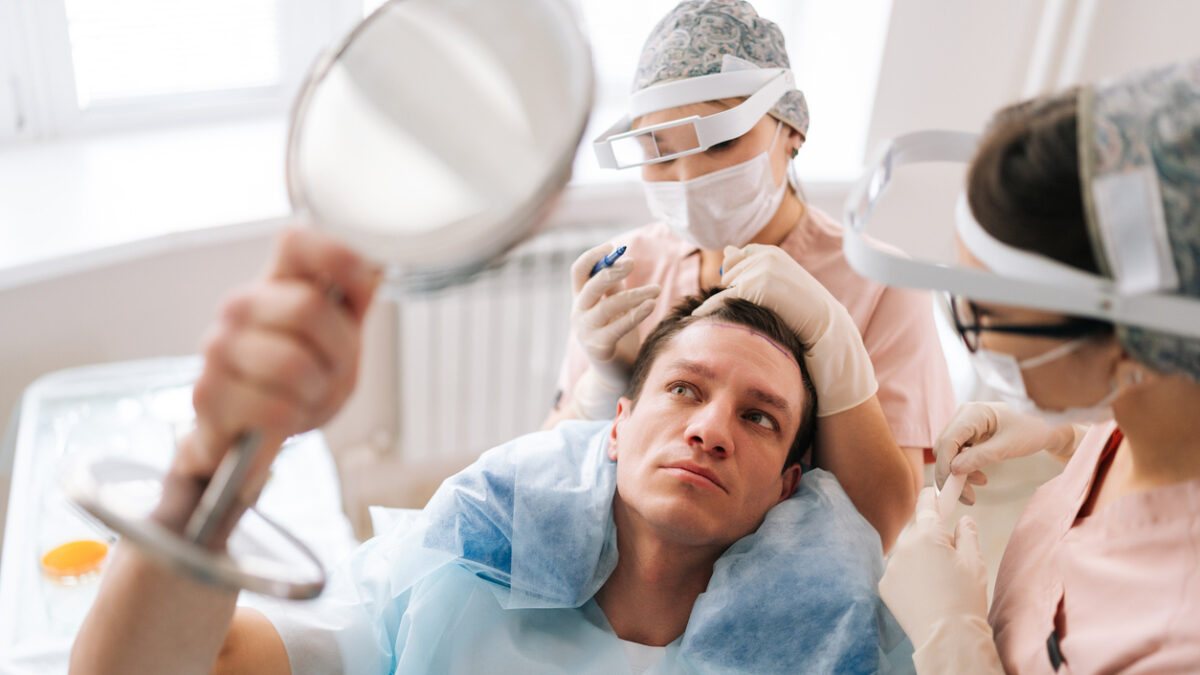 Closeup portrait of male patient with hair loss problem talking with female trichologist marking who with pencil new hairline on head before hair transplantation surgery. Concept of baldness treatment