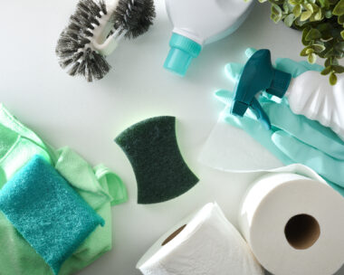 Bathroom cleaning supplies such as disinfectant bottles, scouring pads, gloves, toilet brush and toilet paper on white wooden table. Top view.