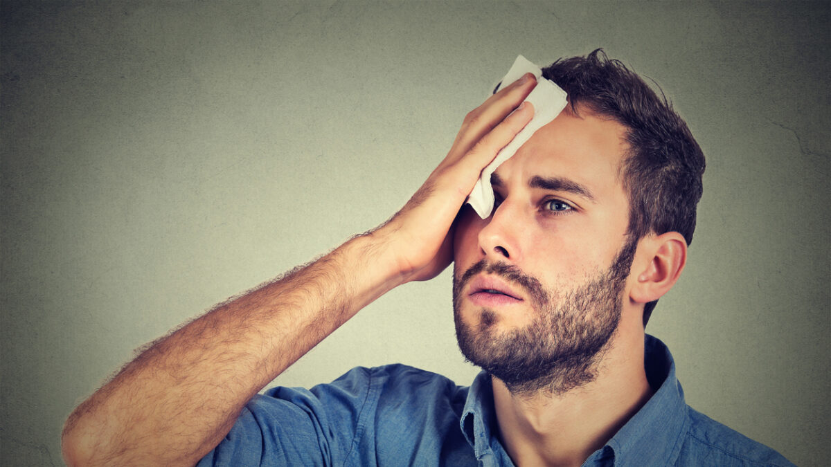 Tired man stressed sweating having fever headache isolated on gray wall background. Worried guy wipes sweat on his face