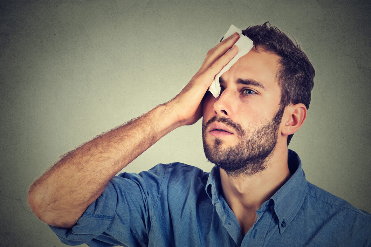 Tired man stressed sweating having fever headache isolated on gray wall background. Worried guy wipes sweat on his face
