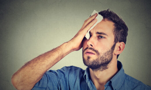 Tired man stressed sweating having fever headache isolated on gray wall background. Worried guy wipes sweat on his face