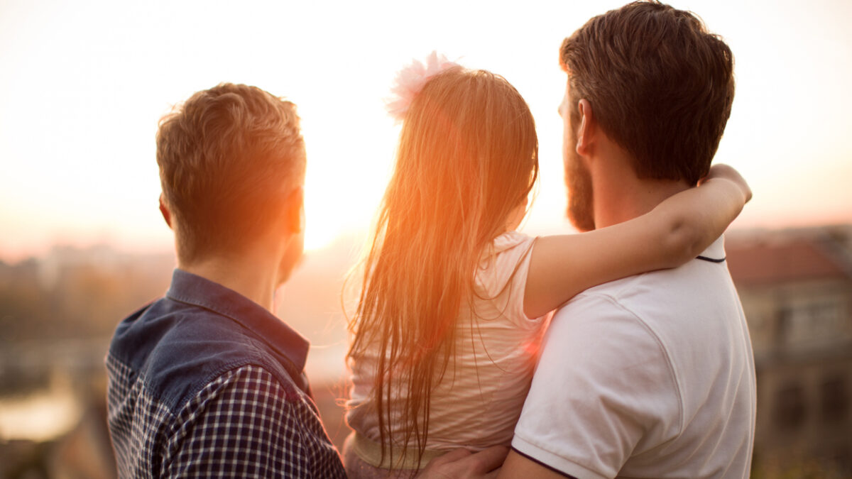 Young gay parents with their daughter having fun in park. Parents holding girl in arms. Enjoying in beautiful sunset. Caucasian ethnicity