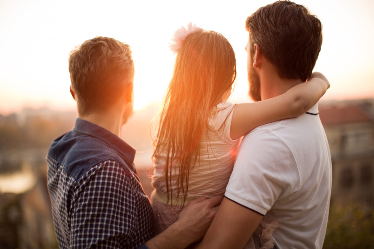 Young gay parents with their daughter having fun in park. Parents holding girl in arms. Enjoying in beautiful sunset. Caucasian ethnicity