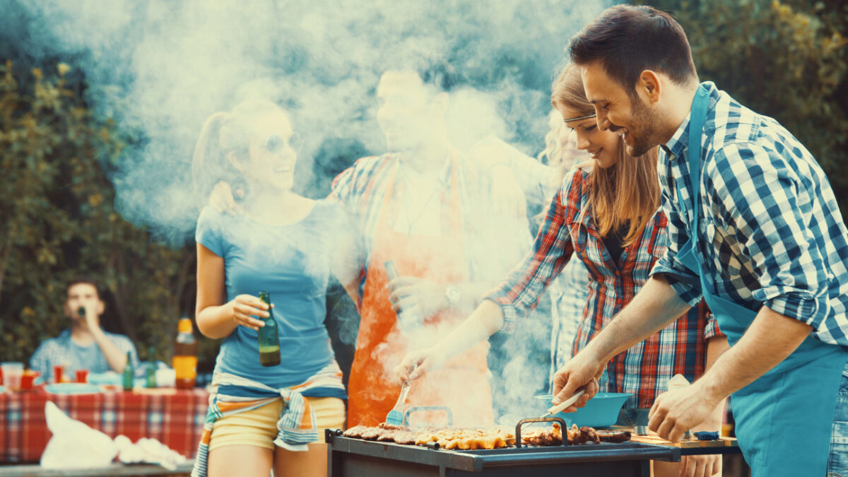 Closeup side view of group of mid 20's people having backyard barbecue party. There are three guys and three girls gathered around heavily smoking grill and sipping cold beer. One of the guys is being today's chef. Toned image, mild contrast