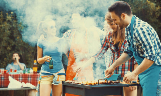 Closeup side view of group of mid 20's people having backyard barbecue party. There are three guys and three girls gathered around heavily smoking grill and sipping cold beer. One of the guys is being today's chef. Toned image, mild contrast