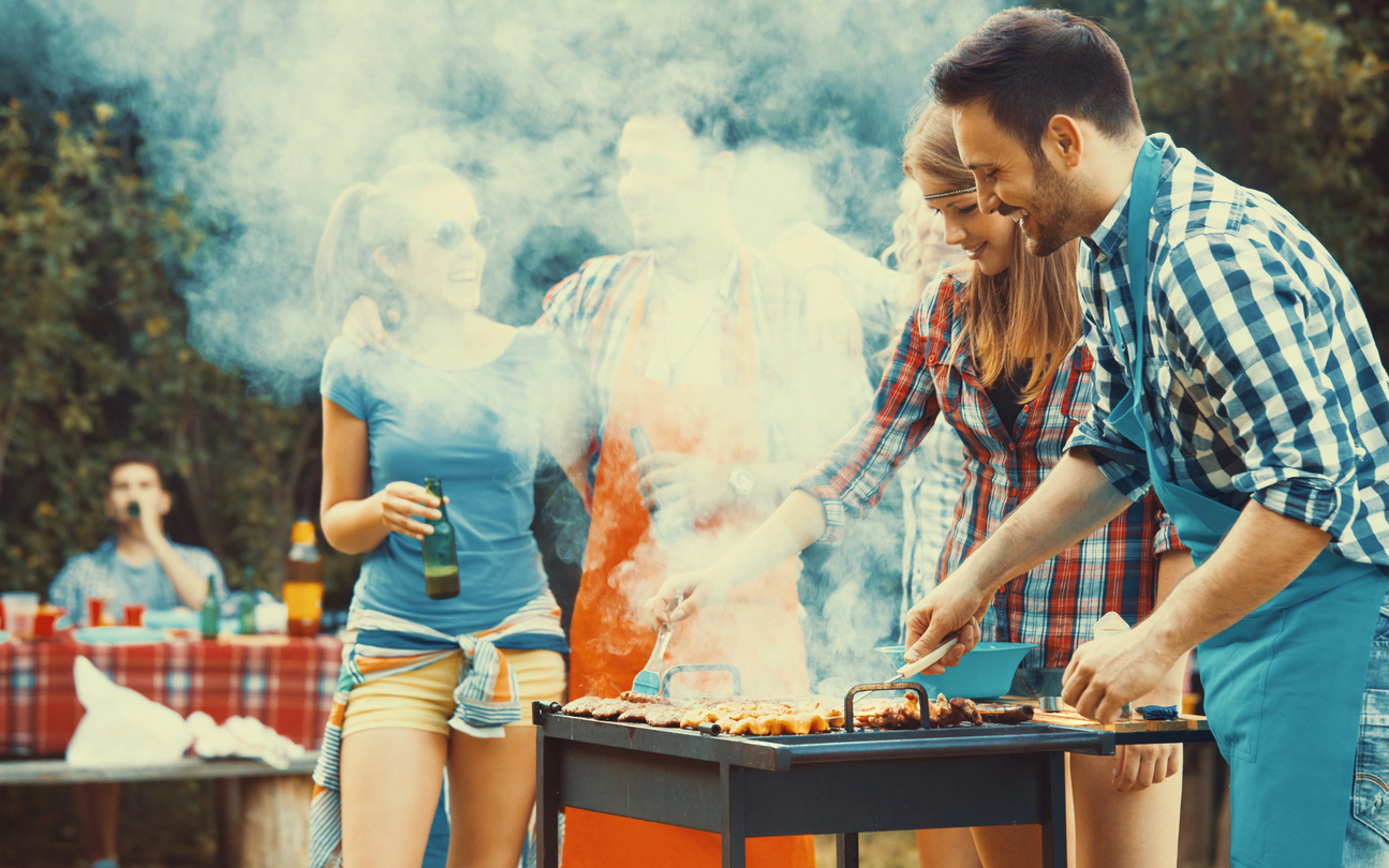 Closeup side view of group of mid 20's people having backyard barbecue party. There are three guys and three girls gathered around heavily smoking grill and sipping cold beer. One of the guys is being today's chef. Toned image, mild contrast