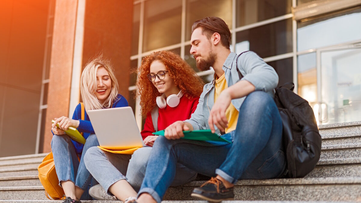 Cheerful young students smiling and doing homework project on laptop while sitting on steps outside university on sunny day
