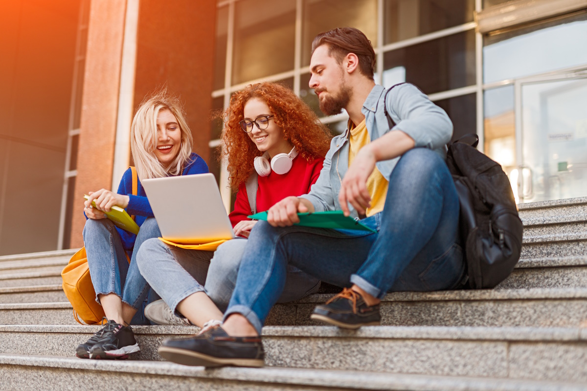 Cheerful young students smiling and doing homework project on laptop while sitting on steps outside university on sunny day