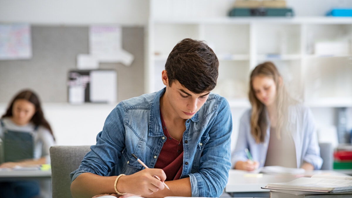 Focused young man taking notes from books for his study. College student sitting at desk with books for finding information in high school library. Guy studying in classroom and completing project with highschool classmates in background.