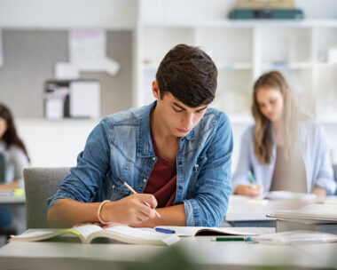 Focused young man taking notes from books for his study. College student sitting at desk with books for finding information in high school library. Guy studying in classroom and completing project with highschool classmates in background.