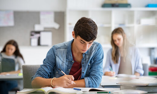 Focused young man taking notes from books for his study. College student sitting at desk with books for finding information in high school library. Guy studying in classroom and completing project with highschool classmates in background.