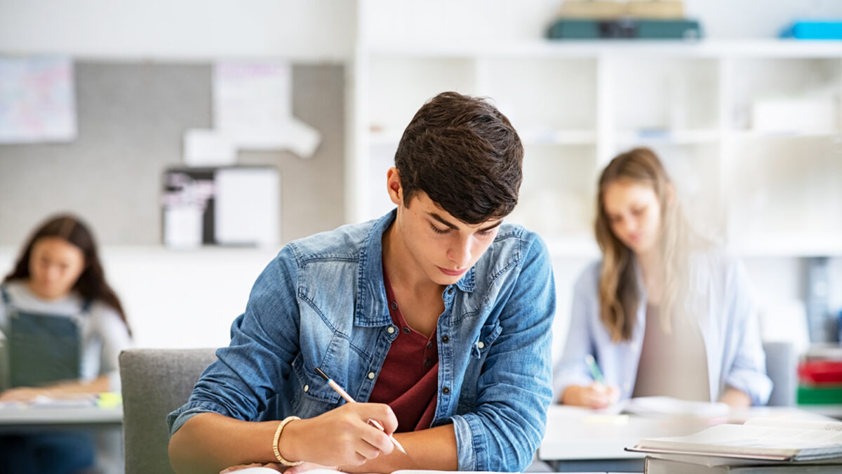 Focused young man taking notes from books for his study. College student sitting at desk with books for finding information in high school library. Guy studying in classroom and completing project with highschool classmates in background.