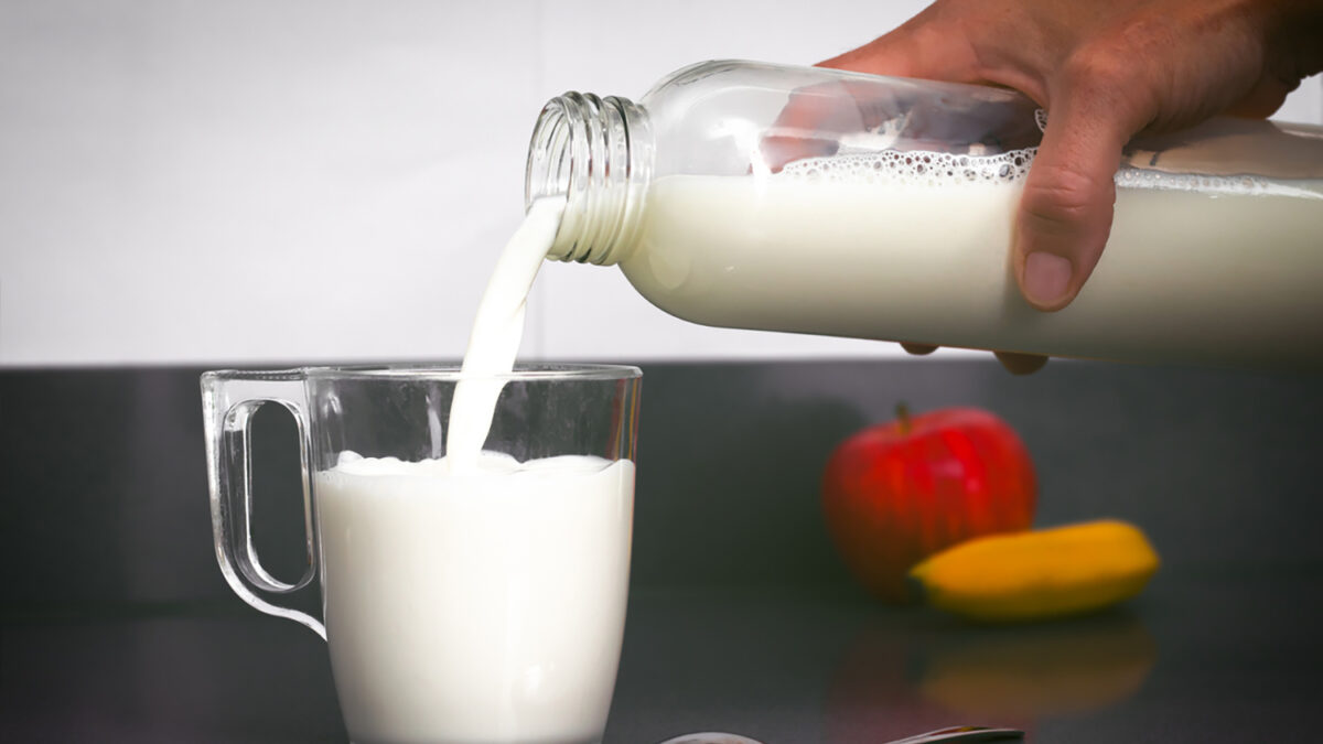 Woman's hand holds a bottle from which milk falls into a small cup, with a teaspoon, a banana and an apple