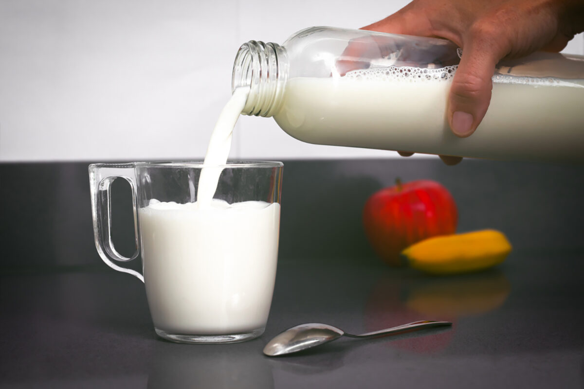 Woman's hand holds a bottle from which milk falls into a small cup, with a teaspoon, a banana and an apple