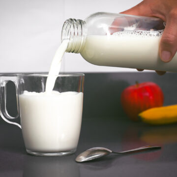 Woman's hand holds a bottle from which milk falls into a small cup, with a teaspoon, a banana and an apple
