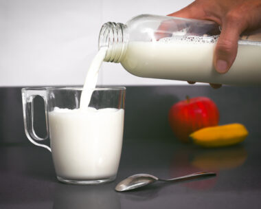 Woman's hand holds a bottle from which milk falls into a small cup, with a teaspoon, a banana and an apple