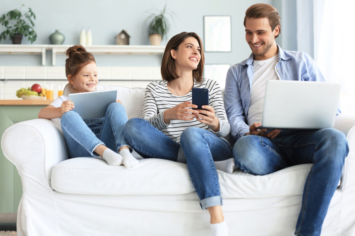 Father, mother and daughter using electronic devices sitting on sofa at living room