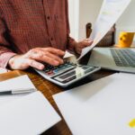 Wide angle image depicting a senior man's hand holding an energy bill while the other hand checks the numbers on a calculator. The table is strewn with documents and also a laptop. Room for copy space.