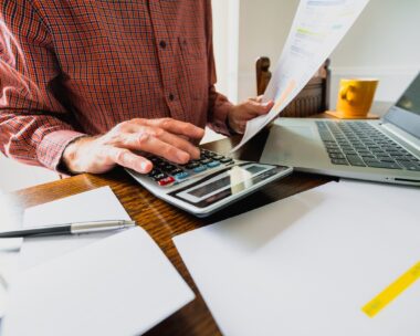 Wide angle image depicting a senior man's hand holding an energy bill while the other hand checks the numbers on a calculator. The table is strewn with documents and also a laptop. Room for copy space.