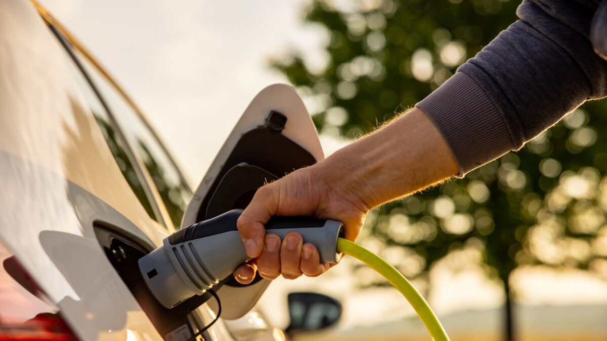 Hand of man inserting a power cord into an electric car for charging ecofriendly vehicle on green landscape
