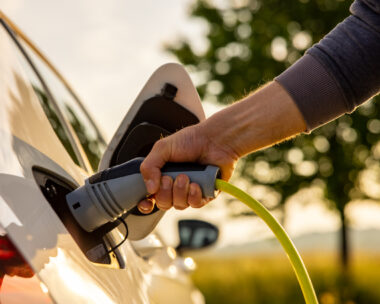Hand of man inserting a power cord into an electric car for charging ecofriendly vehicle on green landscape