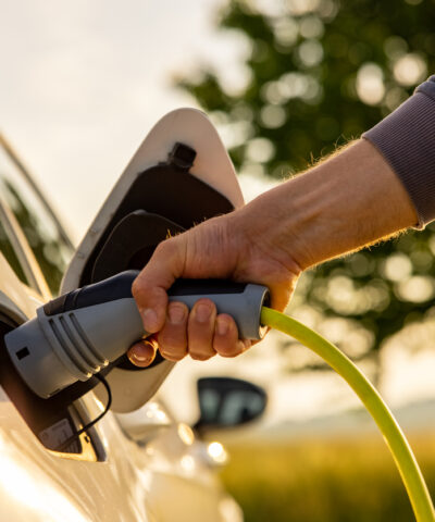 Hand of man inserting a power cord into an electric car for charging ecofriendly vehicle on green landscape