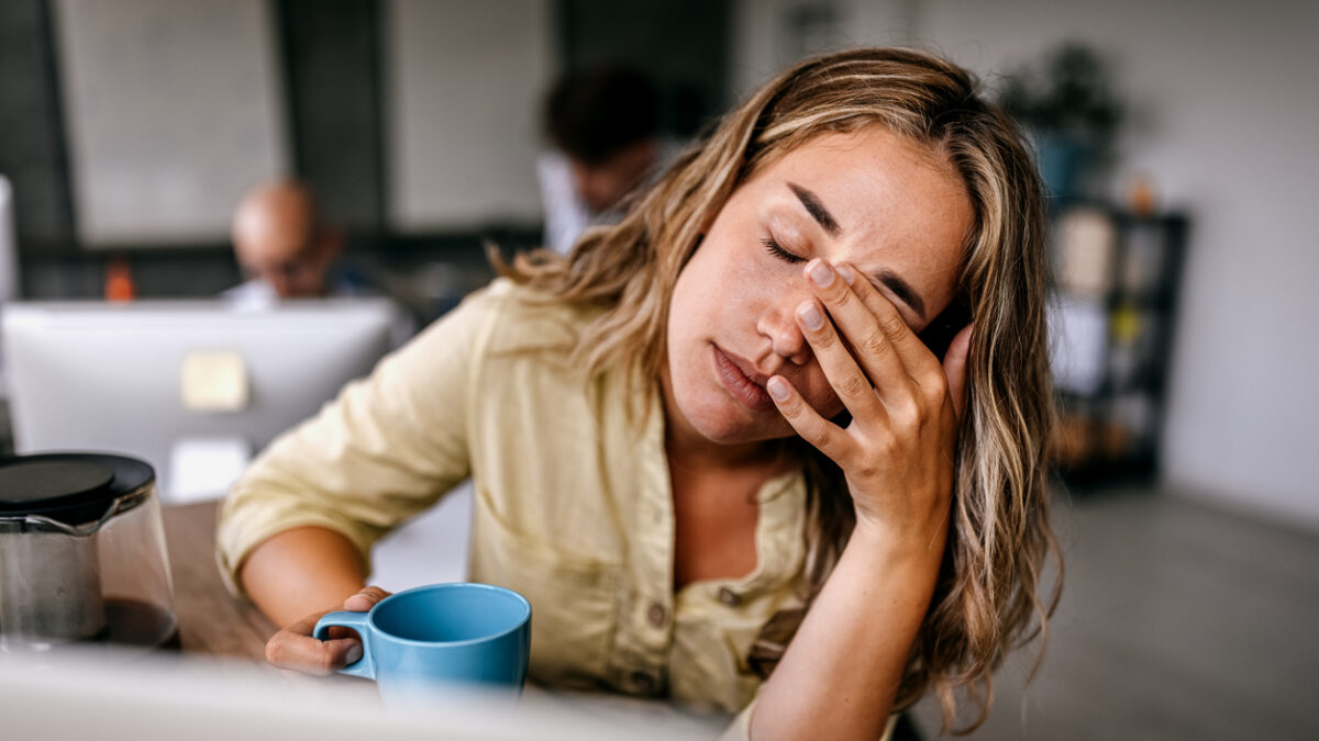 Tired and exhausted young female entrepreneur drinking coffee and rubbing eyes while feeling stressed and worried in coworking space