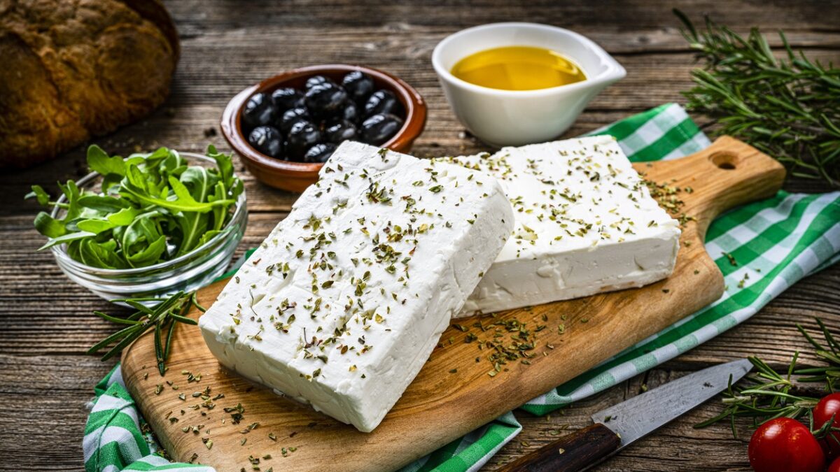 close up view of two blocks of Greek feta cheese on a cutting board shot on rustic wooden table. Olives, cherry tomatoes, olive oil, salt and pepper and herbs like rosemary, oregano, and arugula complete the composition