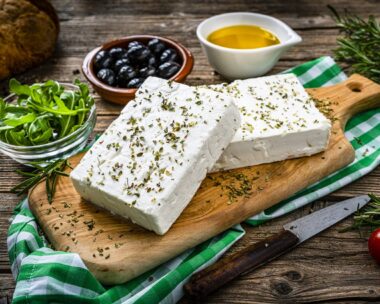 close up view of two blocks of Greek feta cheese on a cutting board shot on rustic wooden table. Olives, cherry tomatoes, olive oil, salt and pepper and herbs like rosemary, oregano, and arugula complete the composition