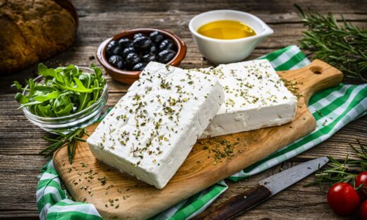 close up view of two blocks of Greek feta cheese on a cutting board shot on rustic wooden table. Olives, cherry tomatoes, olive oil, salt and pepper and herbs like rosemary, oregano, and arugula complete the composition