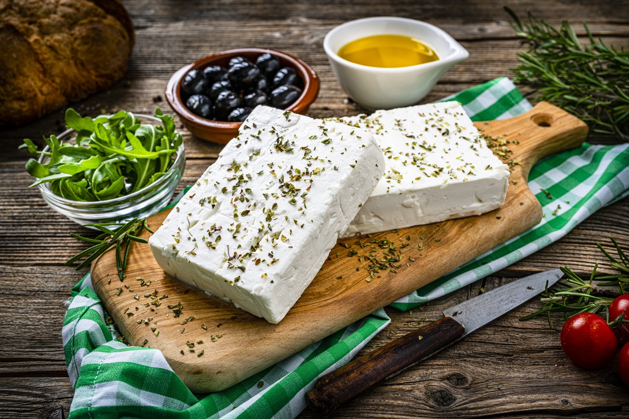 close up view of two blocks of Greek feta cheese on a cutting board shot on rustic wooden table. Olives, cherry tomatoes, olive oil, salt and pepper and herbs like rosemary, oregano, and arugula complete the composition