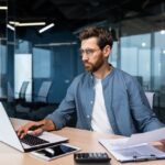 A serious young man accountant, financier, analyst, auditor sits in the office at the table. He holds documents and a pen in his hands, checks accounts, finances, types on a laptop