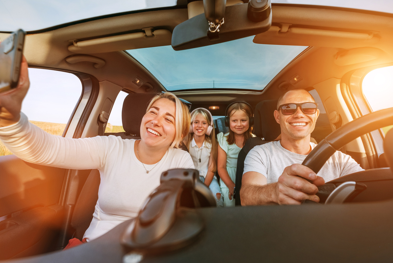 Happy young couple with two daughters inside car during auto trop. They are smiling, laughing and taking selfies using smartphone. Family values, traveling, social media and new technology concepts.