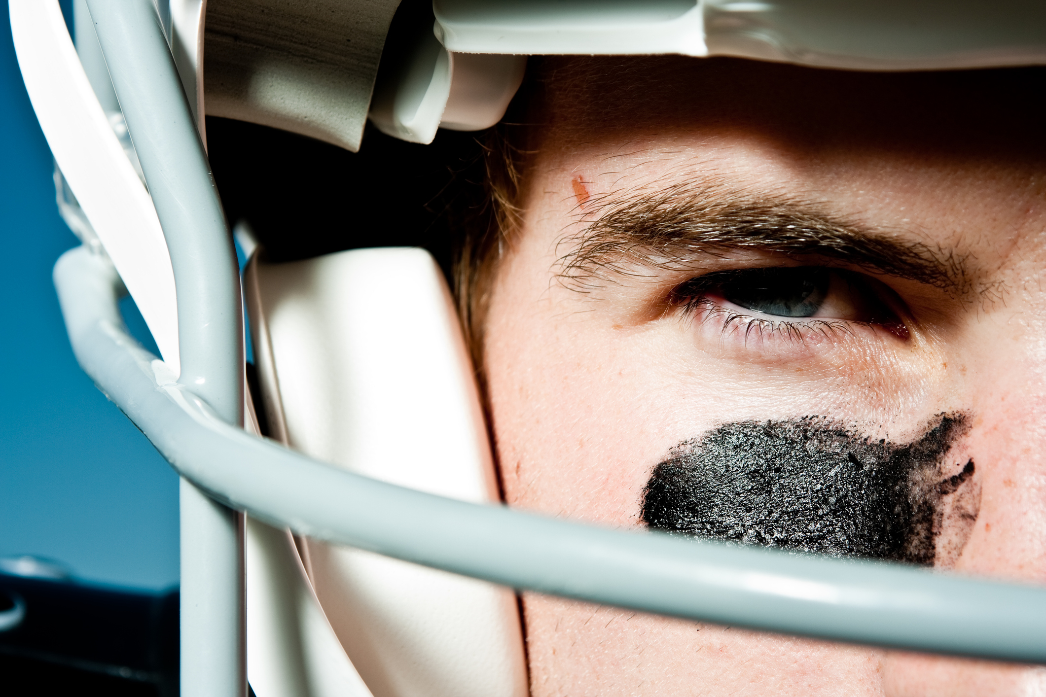 Young Man in his 20s wearing helmet and pads for American style football holding a football. He has eye black on his face and has blue eyes. He has a very serious and competitive look on his face.