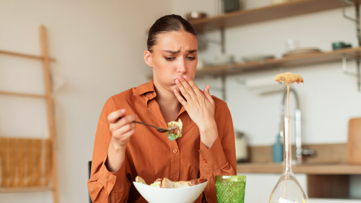 Young caucasian woman feeling nausea while eating delivered caesar salad, lady looking at plate and closing mouth, sitting at table in kitchen interior