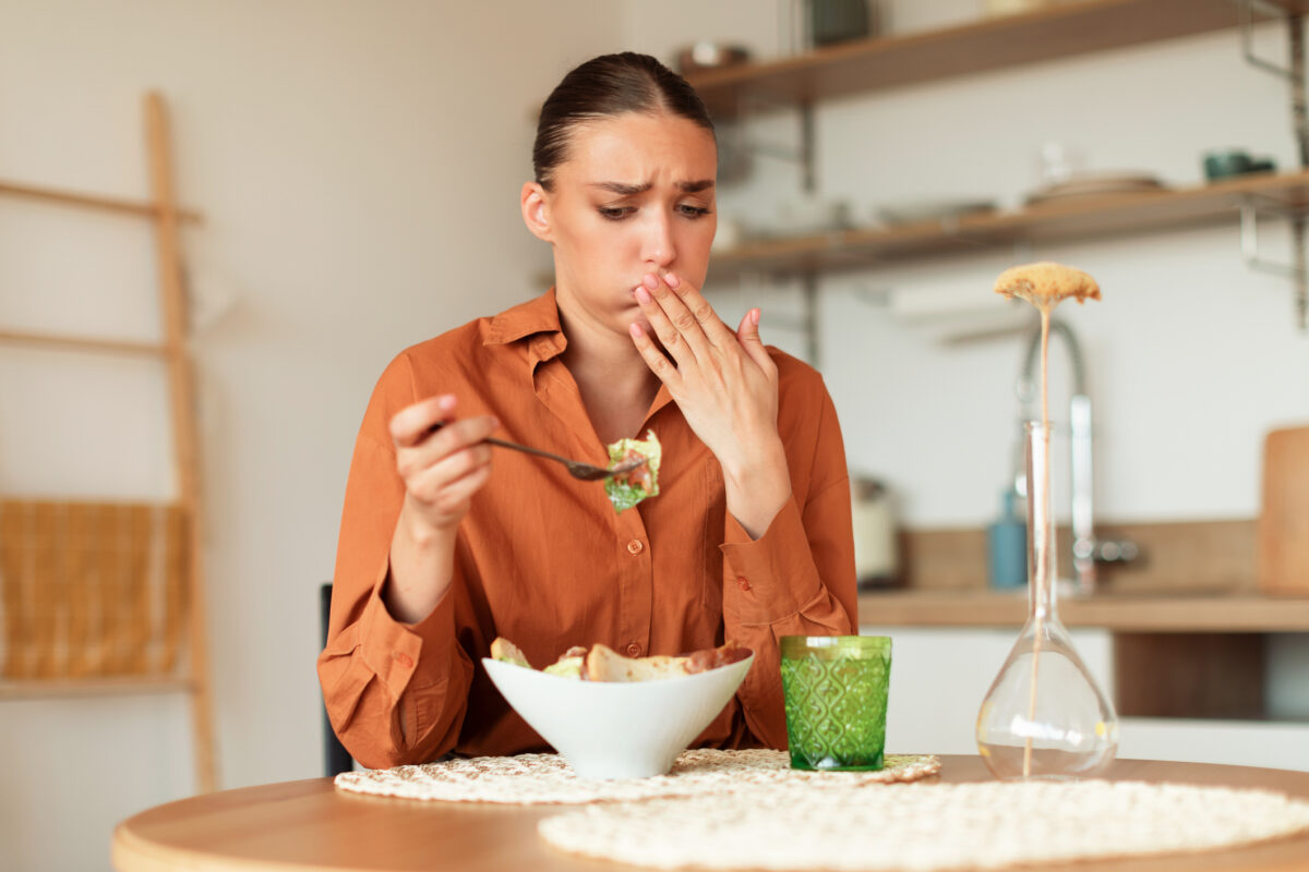 Young caucasian woman feeling nausea while eating delivered caesar salad, lady looking at plate and closing mouth, sitting at table in kitchen interior