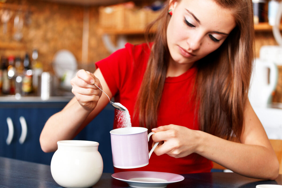 "girl putting white sugar in her tea or coffee - unhealthy lifestyle :)focus on coffee cup & spoon with sugar, the girl & the kitchen is defocused"