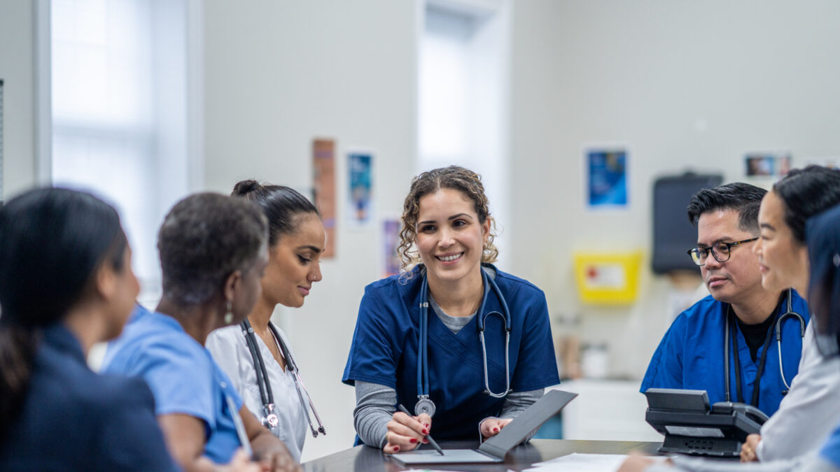 A small group of medical professionals gather around a table as they meet together to discuss patient cases. They are each dressed professionally and have files scattered between them for reference.