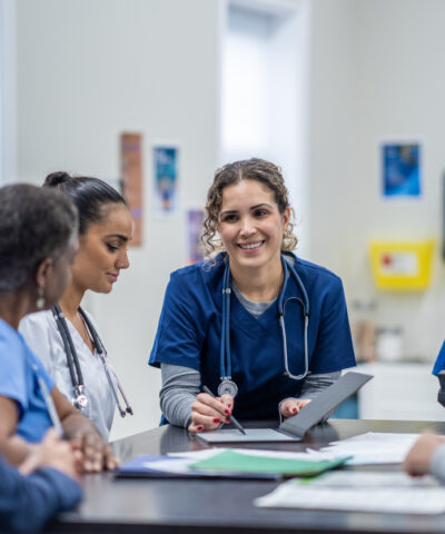 A small group of medical professionals gather around a table as they meet together to discuss patient cases. They are each dressed professionally and have files scattered between them for reference.