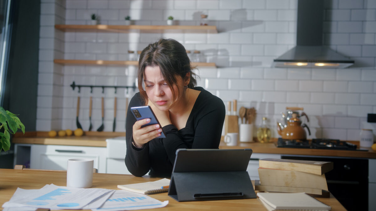 Young woman sit in the kitchen experiences severe pain in neck rubbing to relieve muscle tension massaging neck while using smartphone in kitchen at home